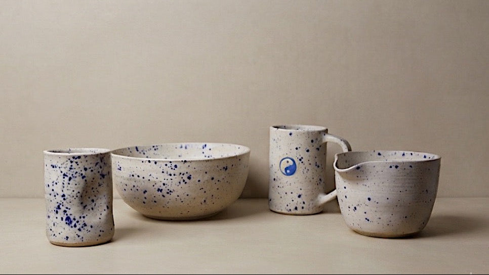 Four handmade ceramic tableware pieces with a speckled glaze, including a bowl, two mugs, and a small pitcher, arranged on a neutral beige surface against a matching background.